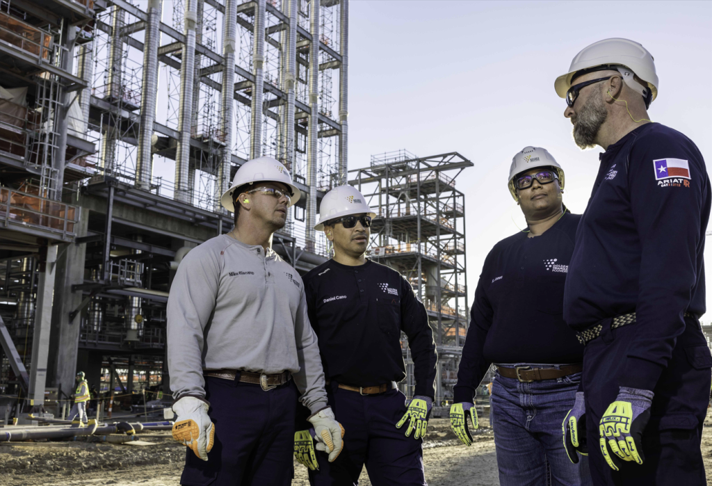 Four people in foreground with white hard hats, with plant in background